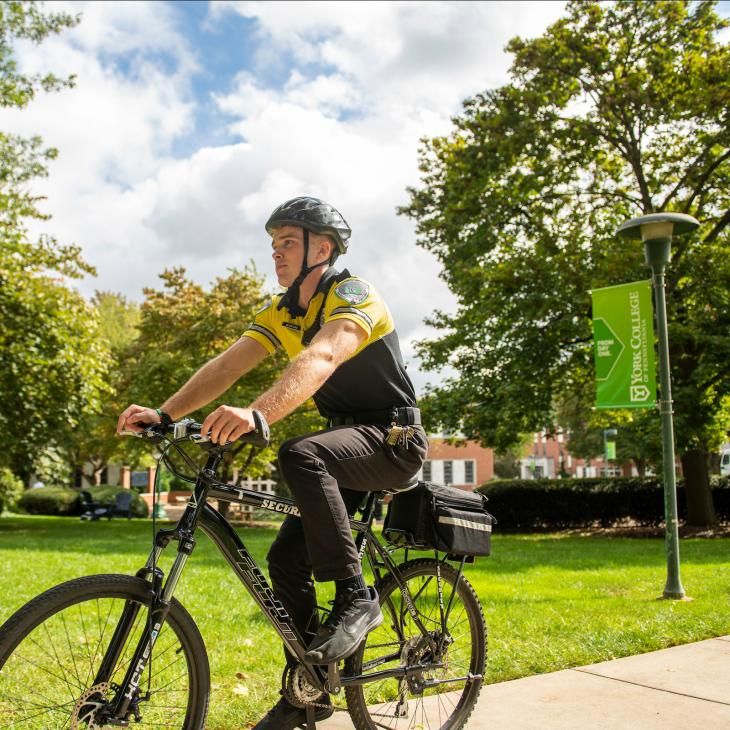 A student security officer wearing a yellow and black uniform rides a bicycle around campus.