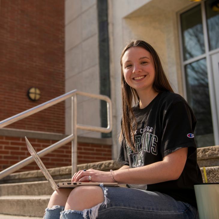 Student sitting on steps outside of the campus library with a laptop on their lap while smiling.