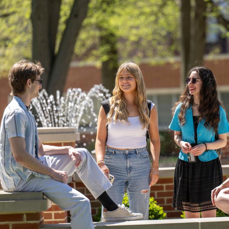 Four students gather near the fountain in the campus quad on a sunny day, chatting and smiling.