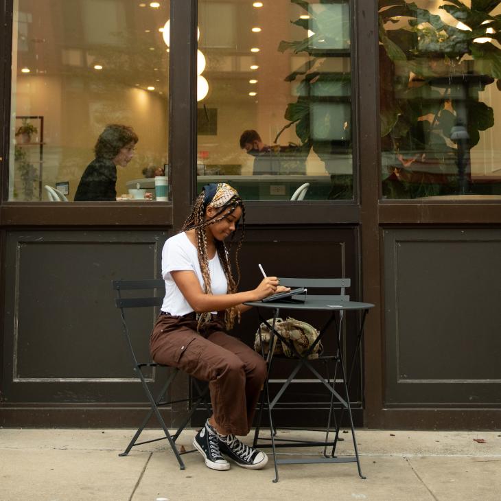 Brianna Simms works on a tablet while sitting at an outdoor table on the sidewalk by a downtown cafe.