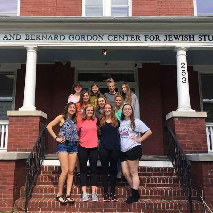 Students standing on the porch and the steps of the Gordon Center. It is a brick building with a white railing around the porch.