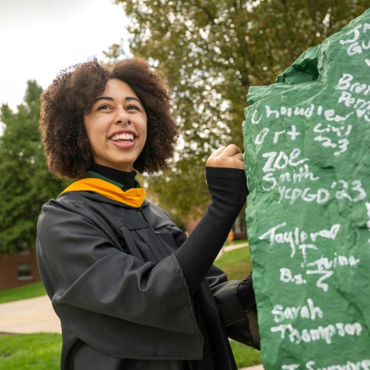 A student in graduation regalia signs the Rock - a green boulder covered in student signatures.
