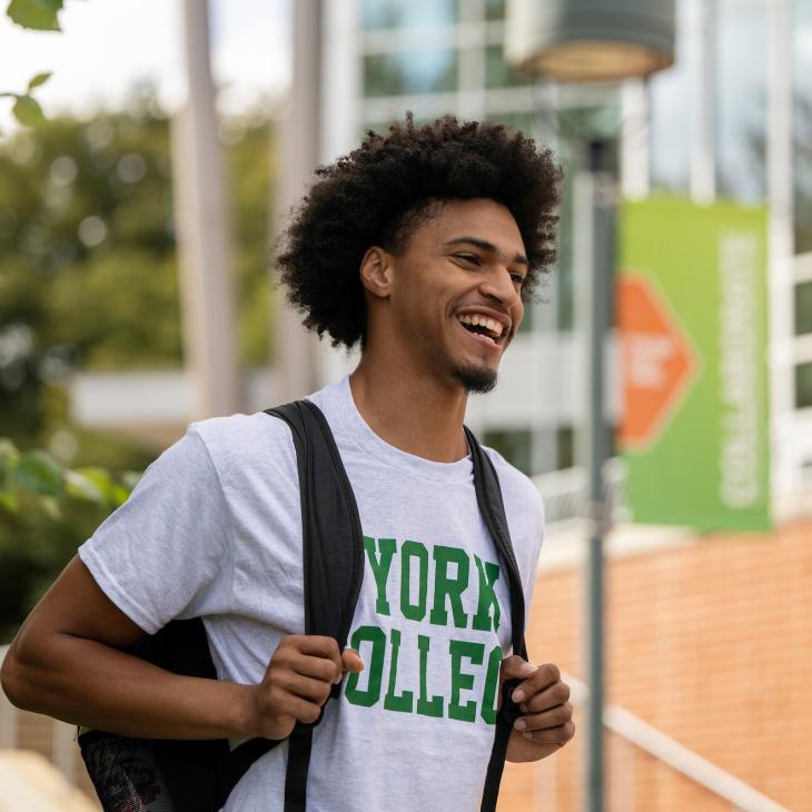 Students walking outside of the Waldner Performing Arts Center while smiling. Student is wearing a white York College tee shirt with green writing.
