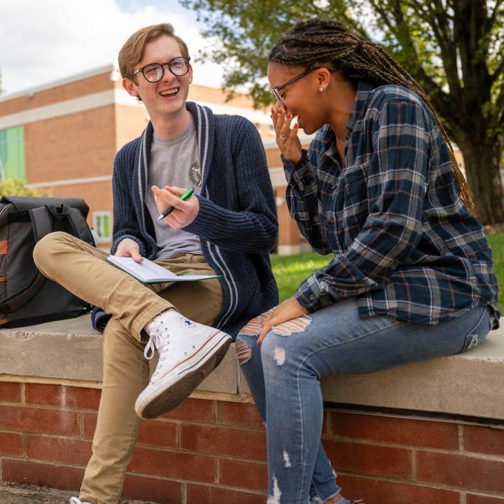 Two students laughing with each other sitting on top of a brick wall