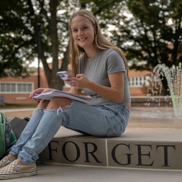 Student sitting on steps in front of the York College fountain taking notes and smiling.