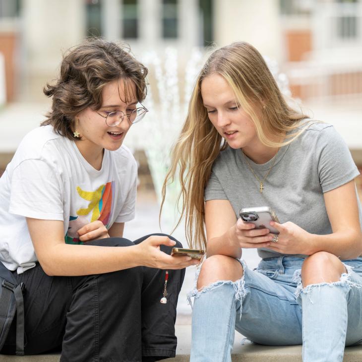Two students sit beside the fountain on Main Campus, glancing at their phones while they chat.