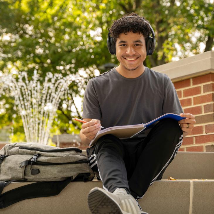 Student sitting on steps outside of the campus fountain. The student is wearing headphones, smiling, and writing in a notebook.
