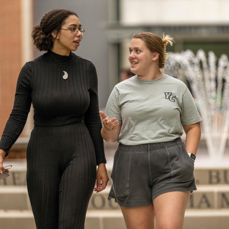 Two students walk on a campus sidewalk. The library and the campus fountain are visible behind them.