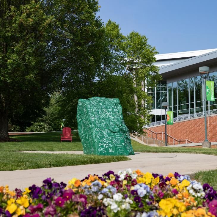 A boulder, painted green and covered in white paint signatures, sits in the center of campus with colorful flowers in the foreground.