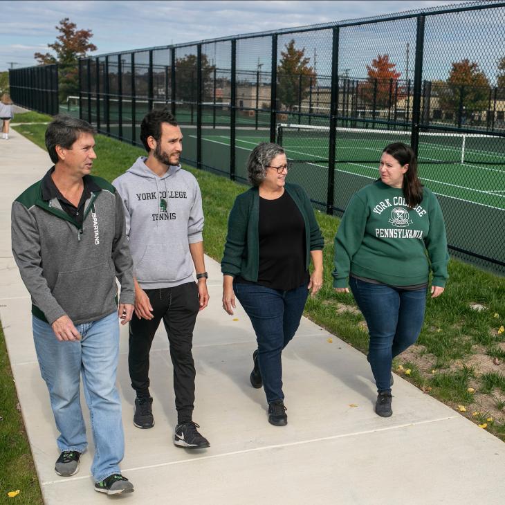 Four people — two traditional-aged college students and two older — walk on the sidewalk along newly painted tennis courts.