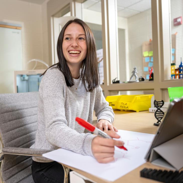 A student writes at a desk, windows and a whiteboard visible behind her, as she looks off-camera and smiles.