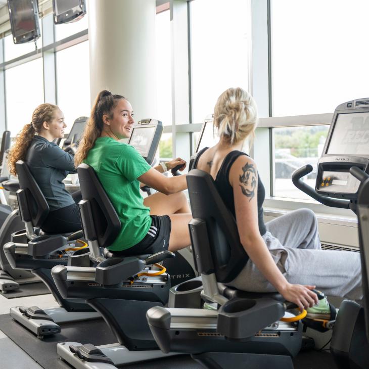 Three students chat while riding recumbent bikes in the fitness center.