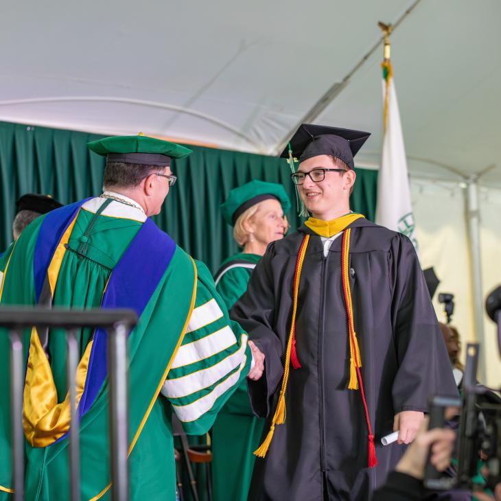 A student in regalia shakes hands with the college president as he crosses the stage at graduation