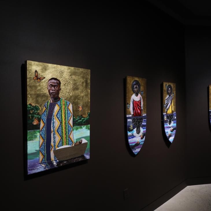 Paintings hang on a black gallery wall, lit dramatically.