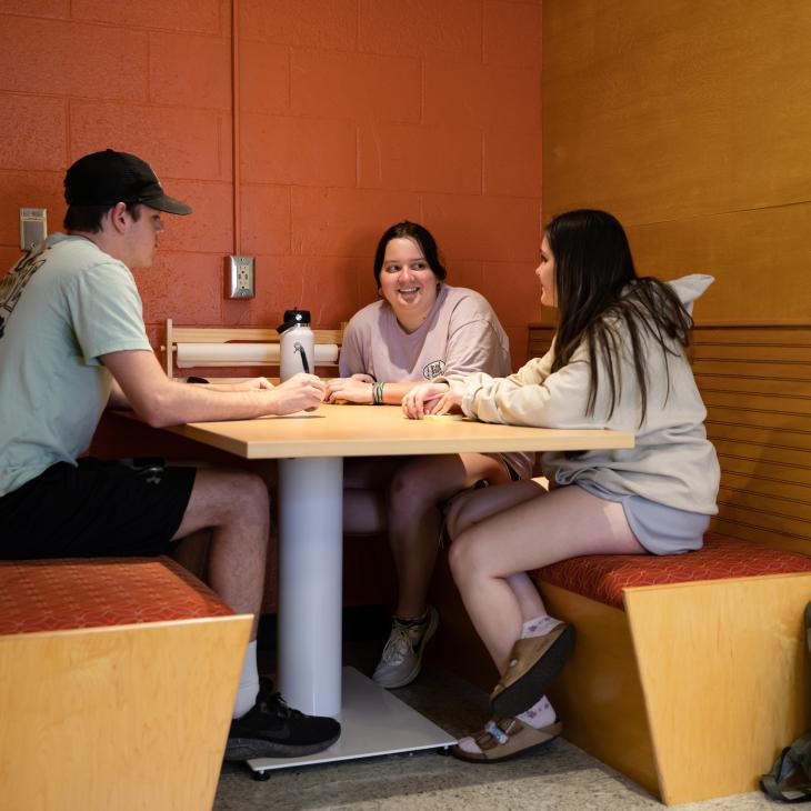 Three students meet at a table in a booth, talking and taking notes.