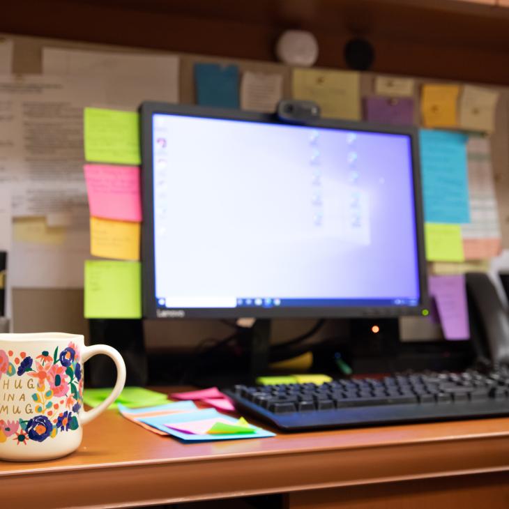 Post-its line the frame of a computer monitor on both sides and are stuck in an organized row on the desk nearby.