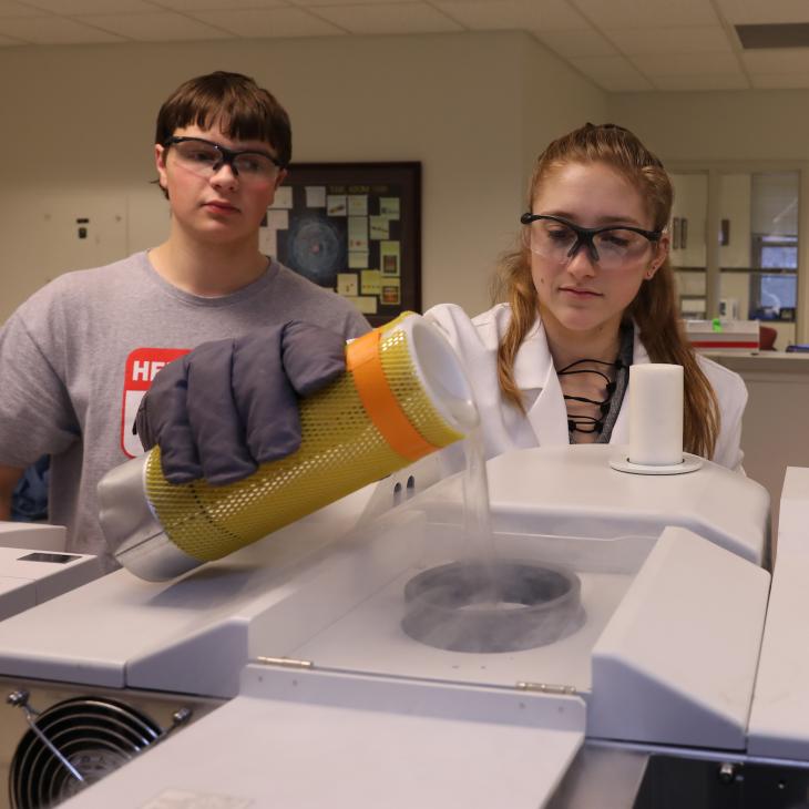 Three students work in the chemistry lab, wearing lab equipment and safety goggles. One pours a chemical into a petri dish while the others look on.