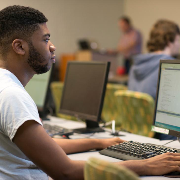 A student works at one of the computers in the computer lab. His classmates are visible in the background, working on their own computers throughout the room.