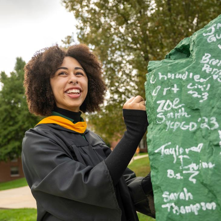 A student wearing a graduation gown writes her signature on a large green boulder.