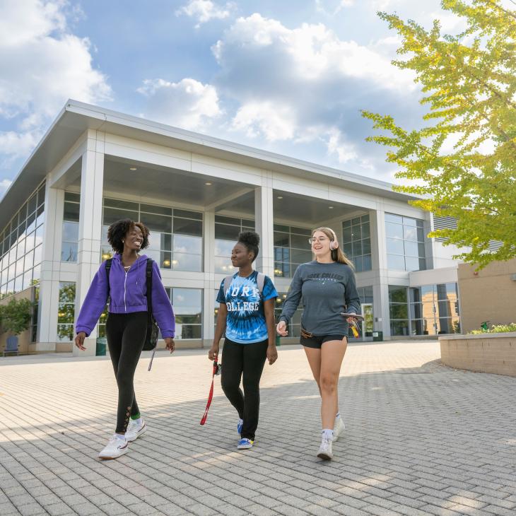 Three students chat and smile as they walk down the sidewalk in front of the Grumbacher Center.