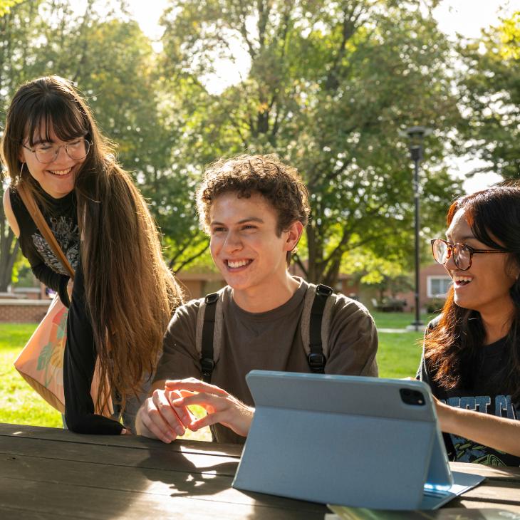 Four students gather outdoors on the campus quad, studying and chatting at a picnic table.