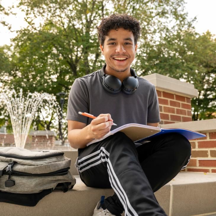 Student sitting at campus fountain with a notebook and pencil