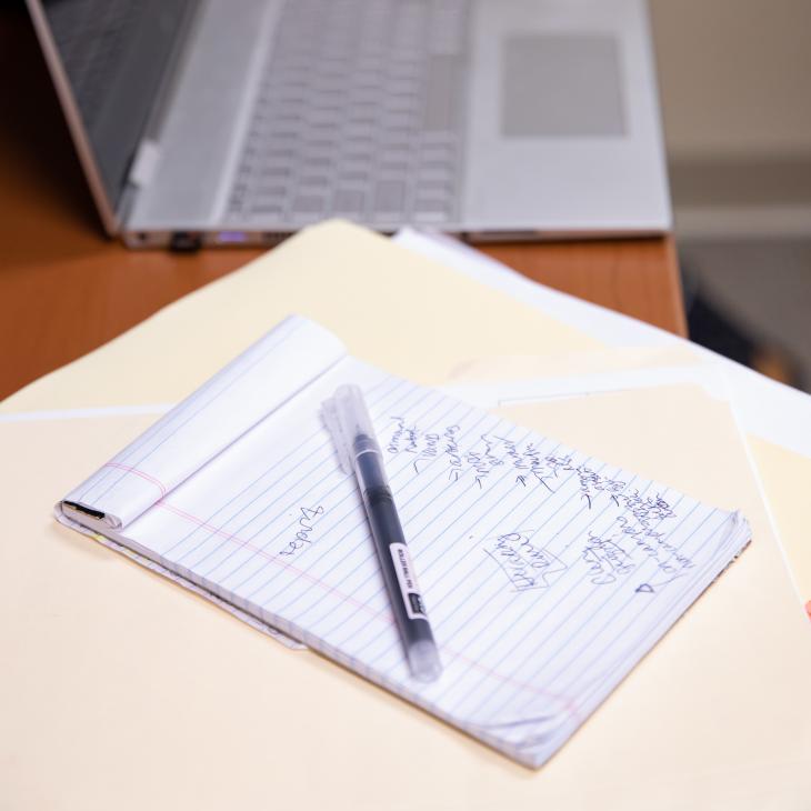 A notepad and pen sit on a desk on top of a manila folder. A laptop is visible in the background.