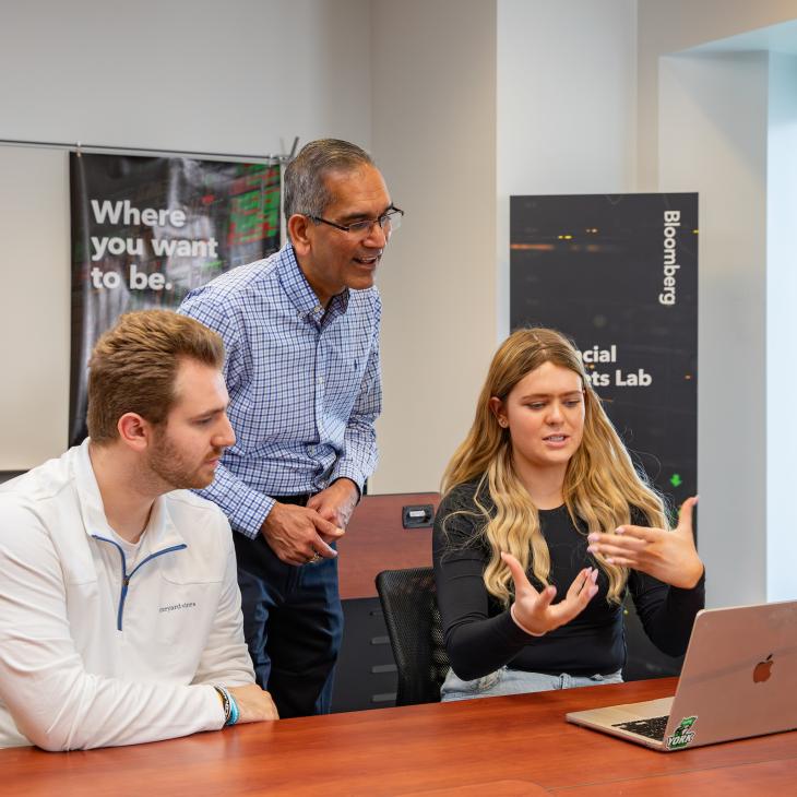 Two students sit at a classroom desk, speaking with a professor as they reference a laptop screen.