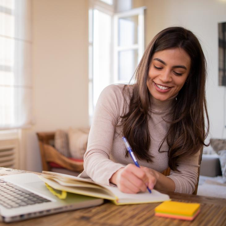 A woman takes notes in a notebook while sitting at a home office desk with her laptop and a pack of post-it notes.