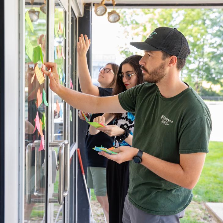 Students attach post-it notes to a window and discuss as part of a design thinking workshop.