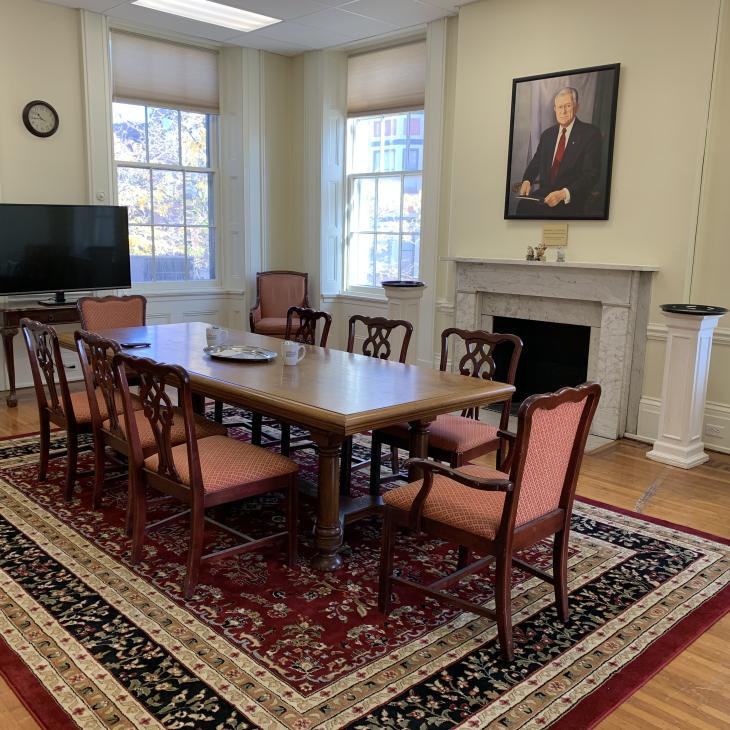 Wooden table and chairs on rug in room with a fireplace, a portait over the mantle, a TV screen, and two tall windows.