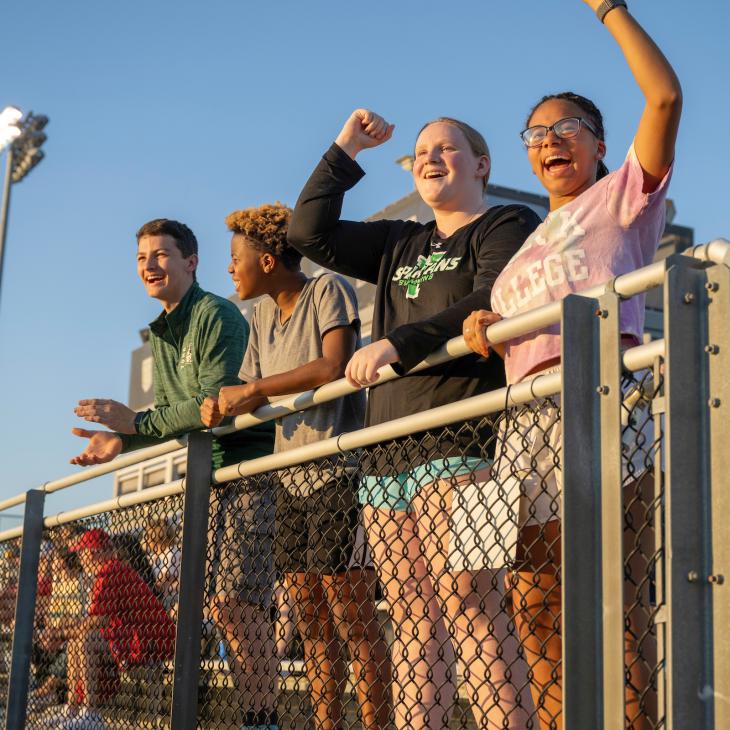 Four students cheer from the bleachers during a York College field hockey game.