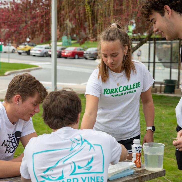 A student in a York College Track and Field t-shirt works on crafts with three other students at an outdoor picnic table on campus.