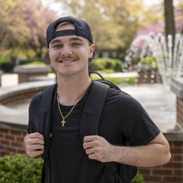 Student wearing a black shirt, hat, and backpack smiles at the camera with an outdoor scene and fountain behind him.