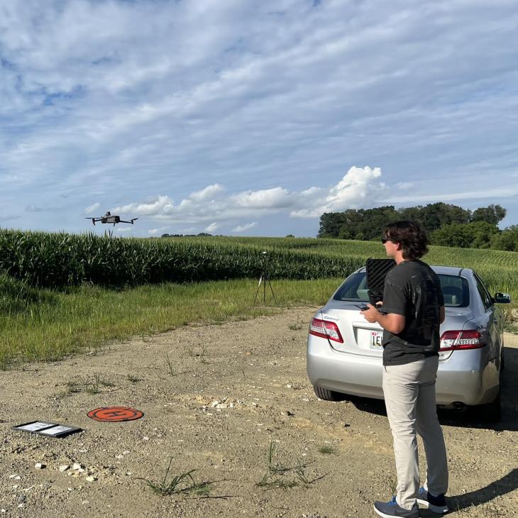 A student operates a drone over a cornfield from a gravel path beside a parked car under a bright blue sky, as part of an outdoor research project.