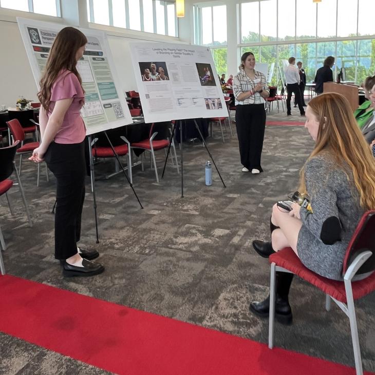 Students present research posters to seated attendees during a formal academic showcase held in a bright, window-lined event space at York College.