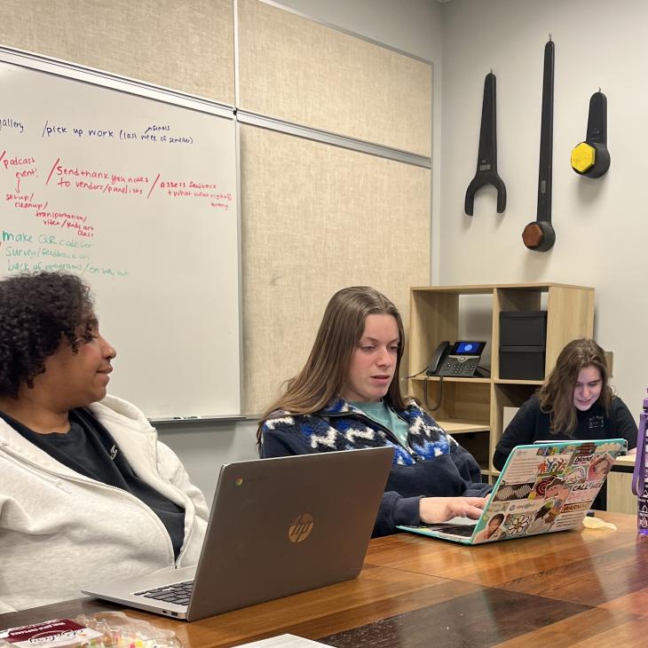 Three students work collaboratively on laptops around a conference table at York College, with notes on a whiteboard and oversized wrenches displayed on the wall behind them.