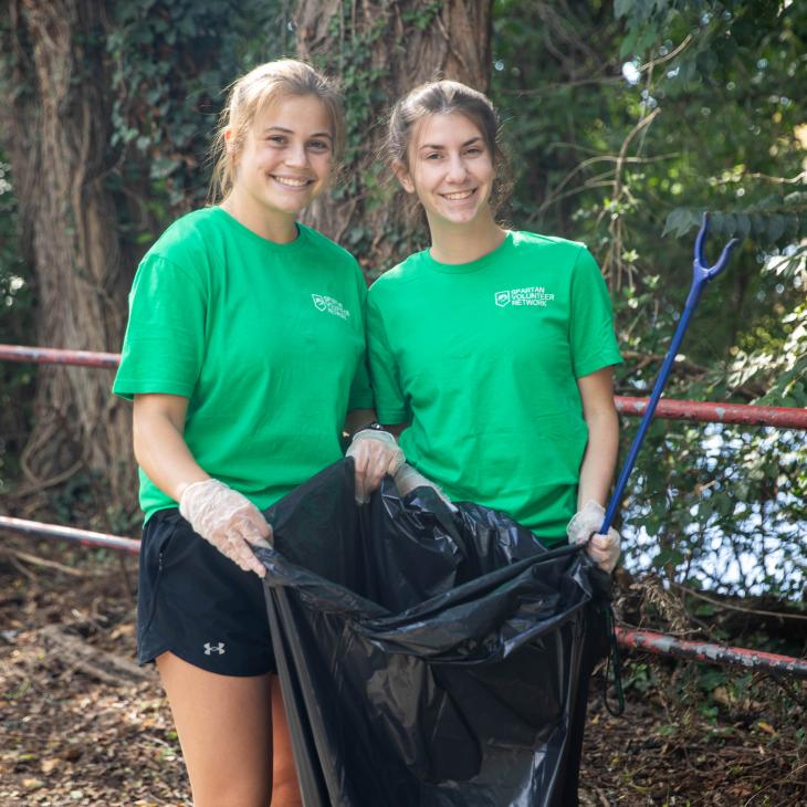 Two students in green t-shirts pose for the camera with a trash bag on the rail trail