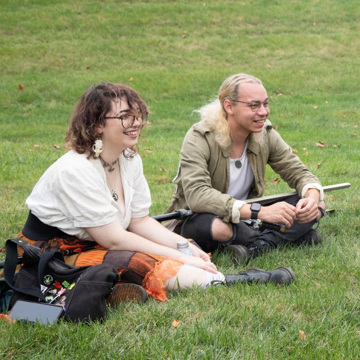 Two students sitting in the grass, they appear to be happily watching an activity out of frame.