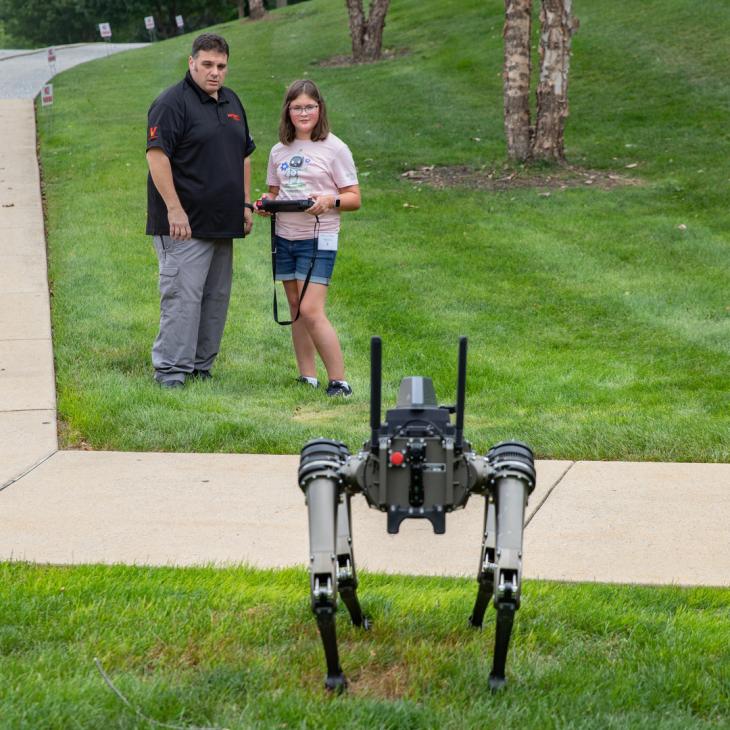 A man stands with a young girl holding a device to control a robot in front of them.