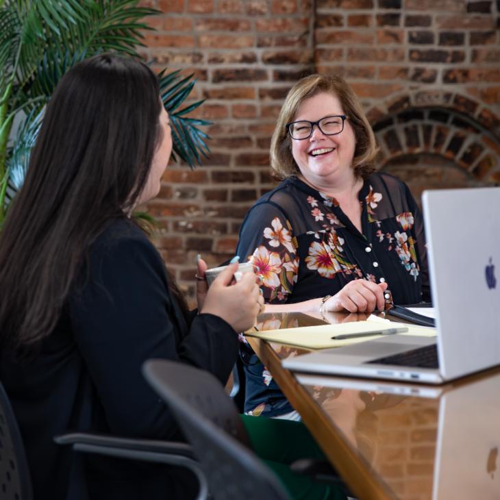 A woman sits at a computer and smiles at another woman. 