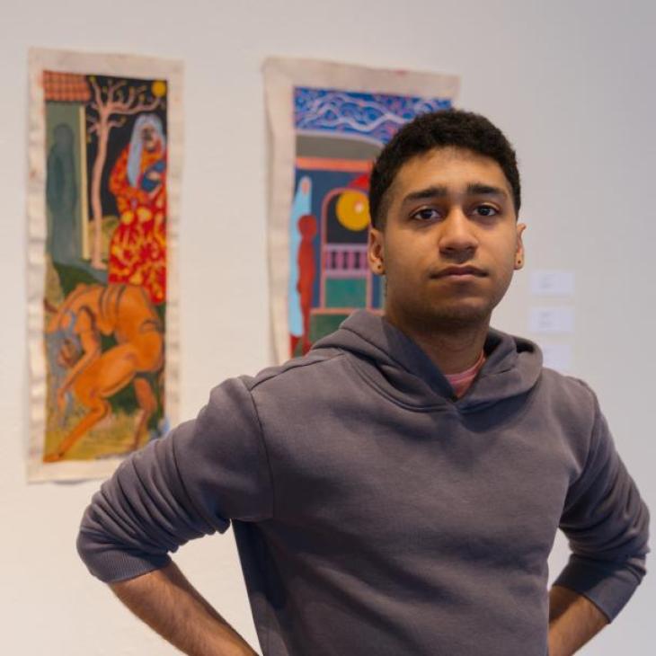2025-26 Appell Arts Fellow Gabriel Sanchez Carpio standing in front of two of his paintings from his solo exhibition, "Cuero."