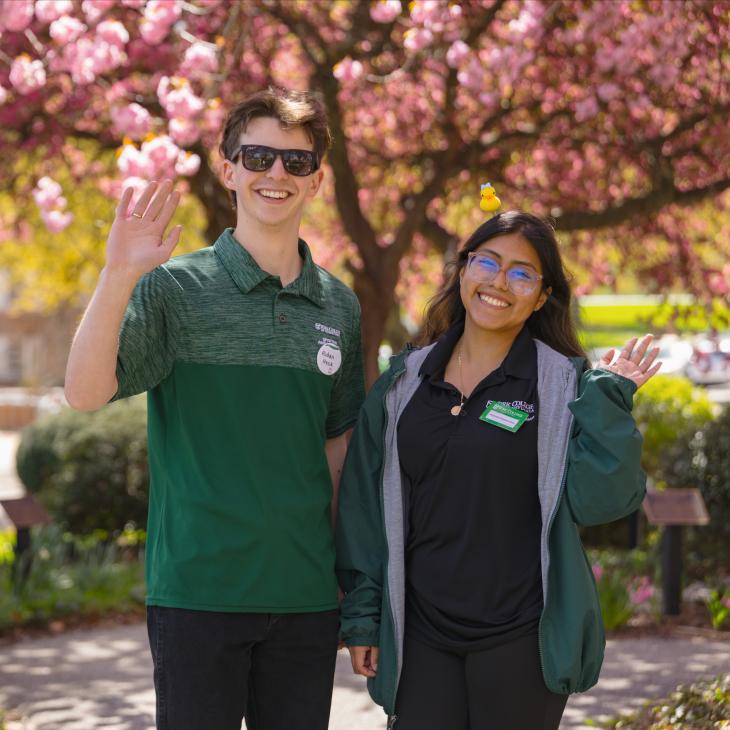 Two student ambassadors smile and wave to the camera with brightly colored flowering trees behind them.