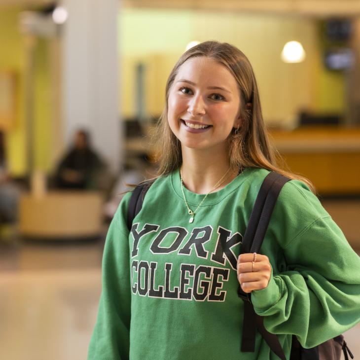 A student wearing a green York College sweatshirt smiles at the camera.